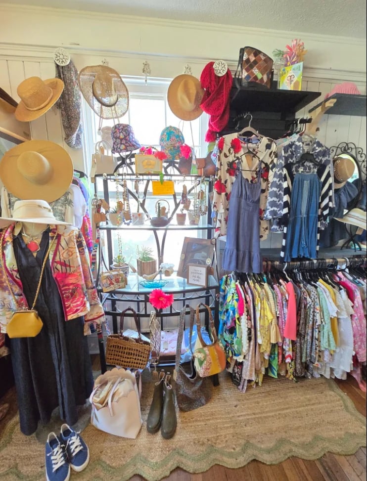 Cluttered retail clothing display with colorful dresses, hats, shoes, and accessories arranged on racks and shelves near a bright window