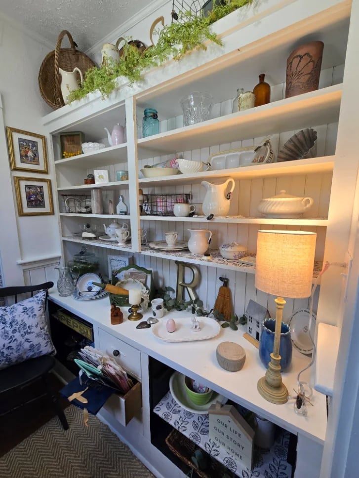 White built-in shelving unit displaying decorative items, plants, books, dishware, and a desk lamp with warm lighting
