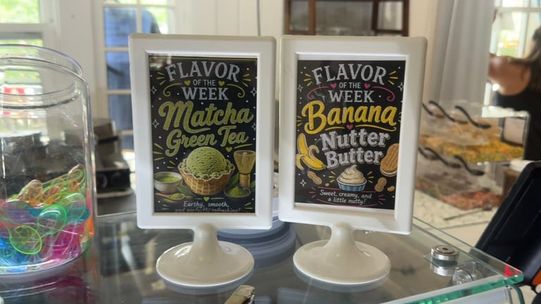 Two black Flavor Week promotional signs displaying Banana Green Tea and Banana Nutter Butter flavors on white stands, displayed on a table with glasses.