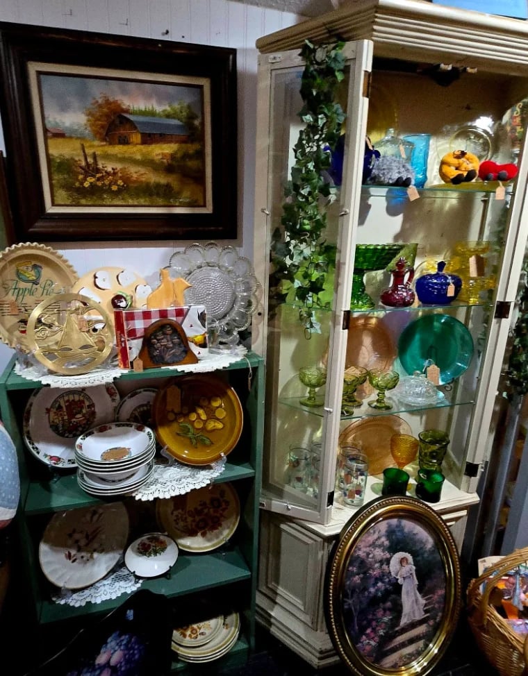 Antique display shelves with vintage farmhouse painting, decorative plates, colorful glass bottles, ceramics, and collectibles arranged in green shelving and glass cabinet