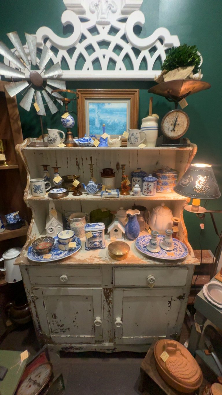 Vintage white cabinet with shelves displaying decorative pottery, china plates, vintage items, and trinkets against a teal wall with ornamental crown