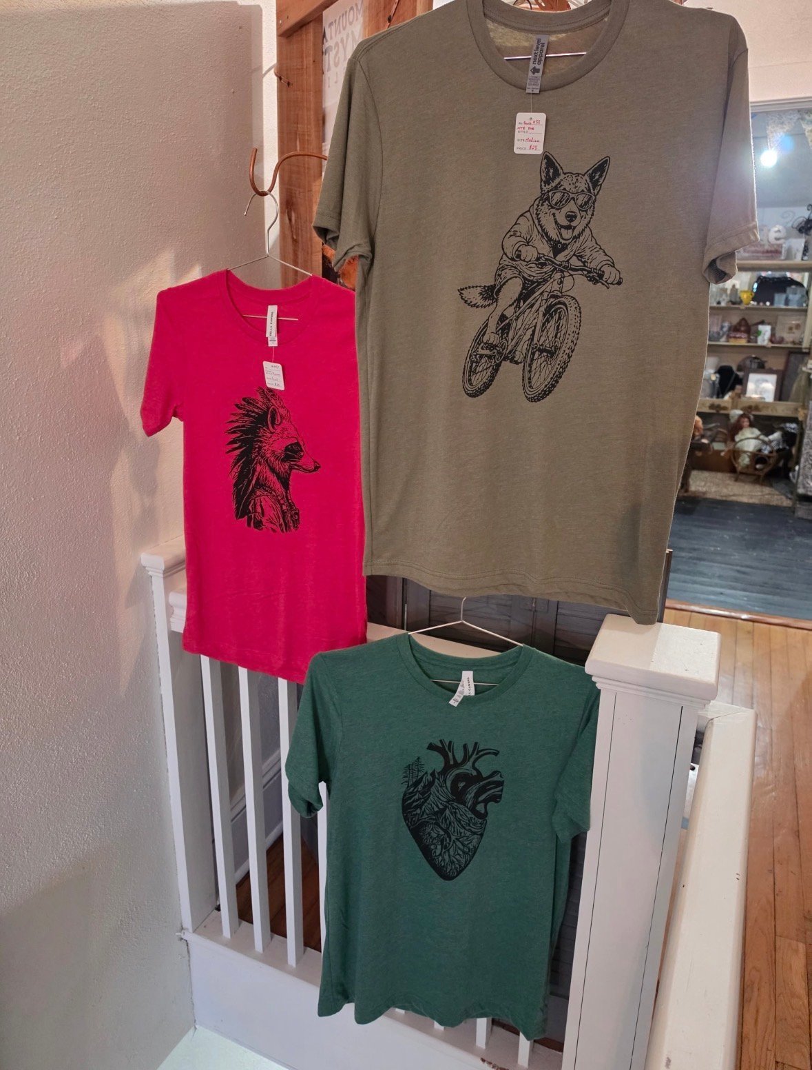 Three graphic t-shirts on display: red with feather design, tan with dog on bicycle, and green with anatomical heart