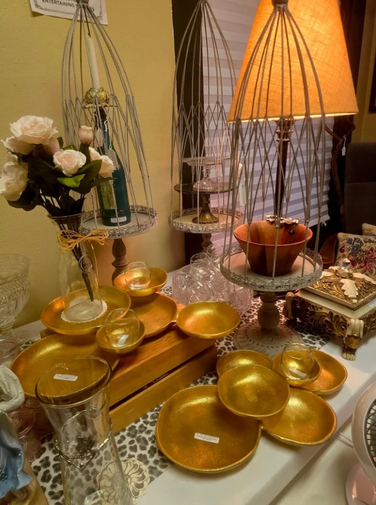 Decorative tabletop display with golden bowls and plates, wire birdcage pendant lamps, white roses in a vase, and home decor items