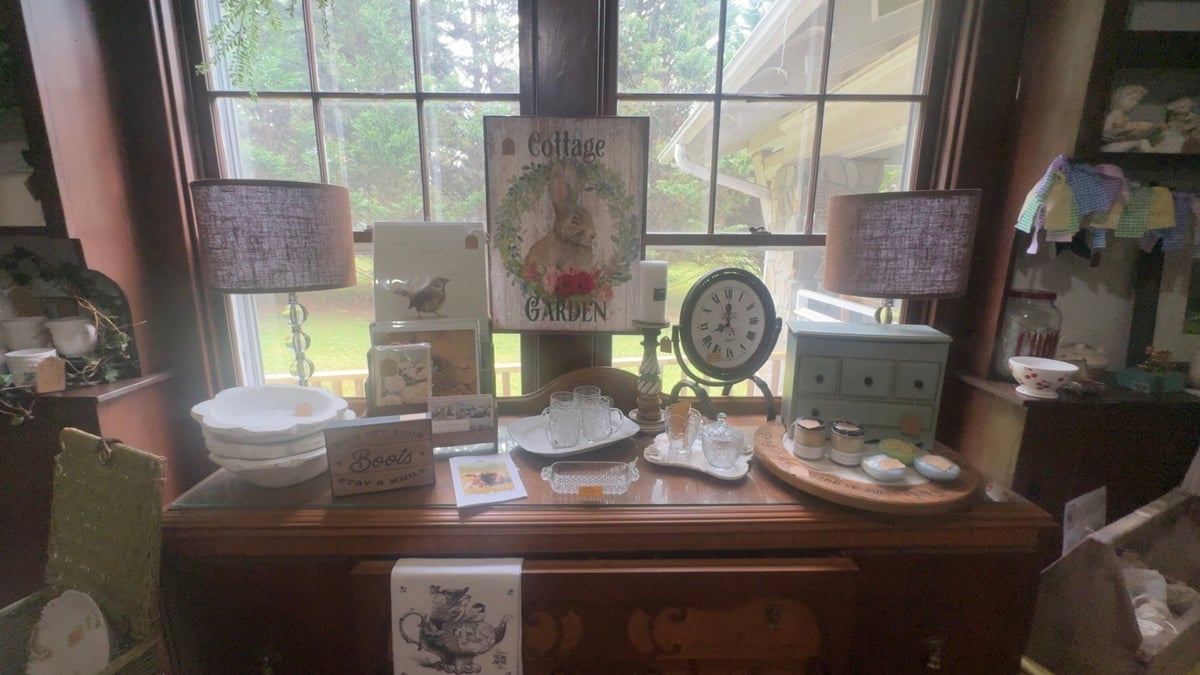 Windowsill display with vintage collectibles including a clock, garden sign, framed pictures, decorative plates, and various ornaments arranged on a wooden shelf.