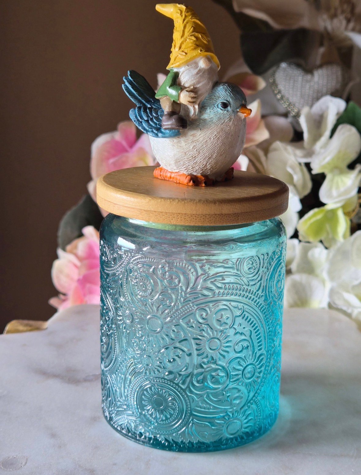 A ceramic bird figurine wearing a yellow hat perches on a wooden lid atop a turquoise embossed glass jar, surrounded by colorful flowers.