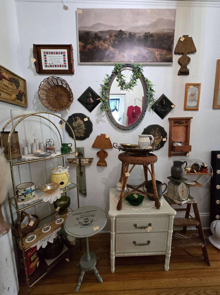 Eclectic bedroom corner with wall-mounted mirrors, framed artwork, decorative items, small furniture pieces, and vintage accessories arranged on shelves and surfaces