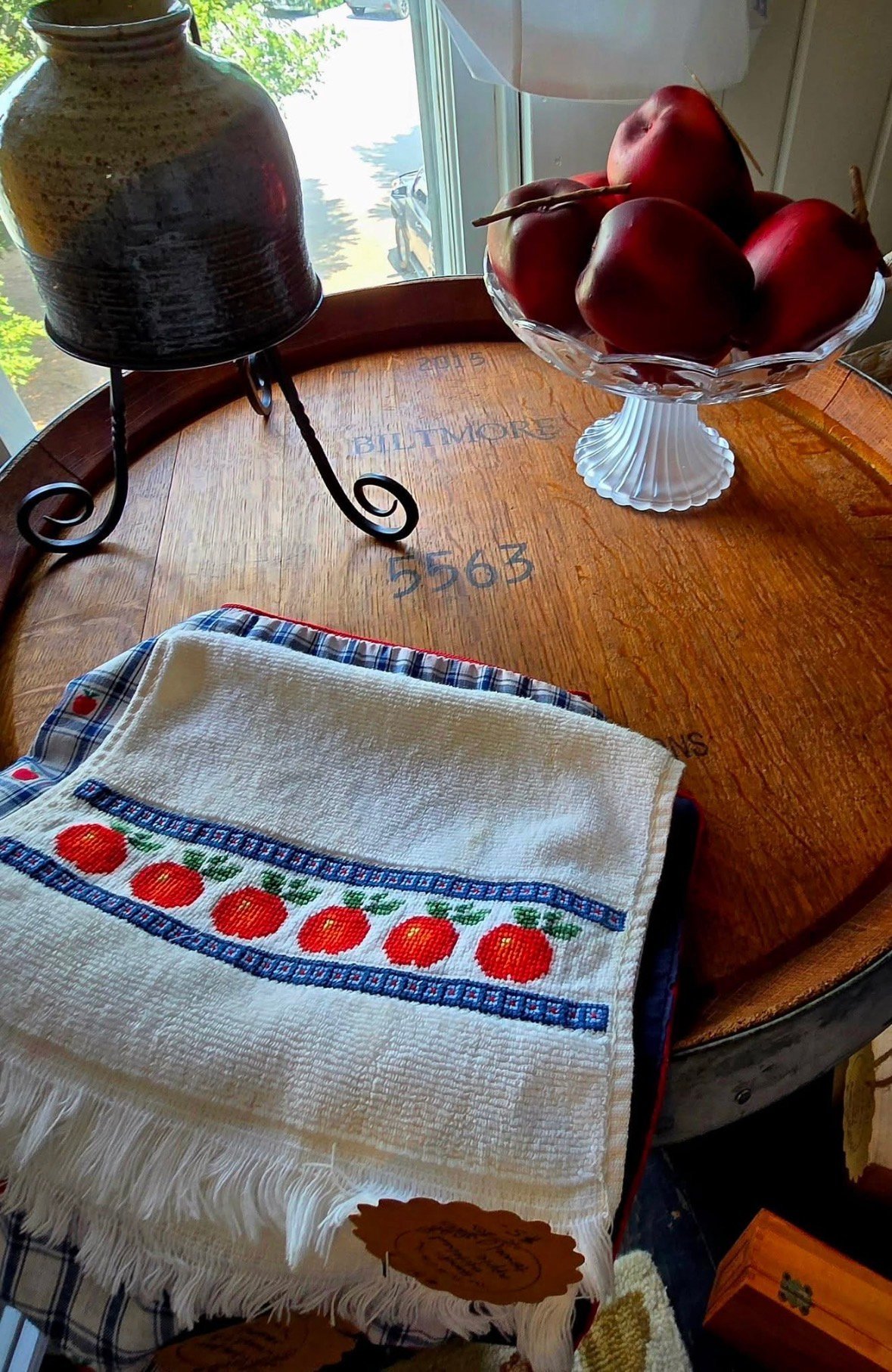 Wooden table with fresh red apples in a pedestal bowl, a kitchen towel with apple motif, a dark glass jar, and a wrought iron stand by a sunny window