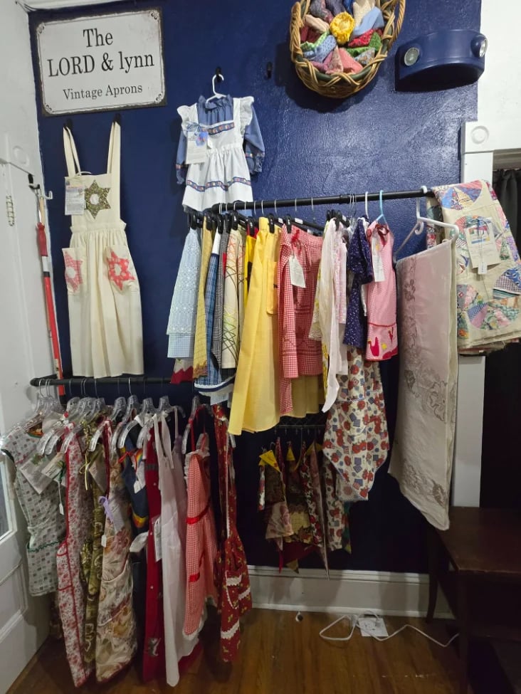 Vintage apron display by The LORD and Lynn brand featuring colorful dresses and aprons hanging on dark blue wall with floral basket decoration