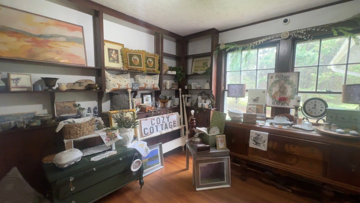 A vintage antique shop interior displaying collectibles, framed artwork, shelves, and furniture against wooden paneled walls with windows