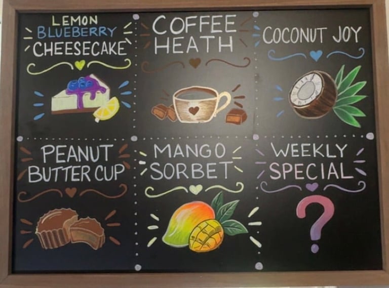 Cafe menu board with chalk art showing six coffee and dessert options including lemon blueberry cheesecake, coffee heath, coconut joy, peanut butter cup, mango sorbet, and weekly special