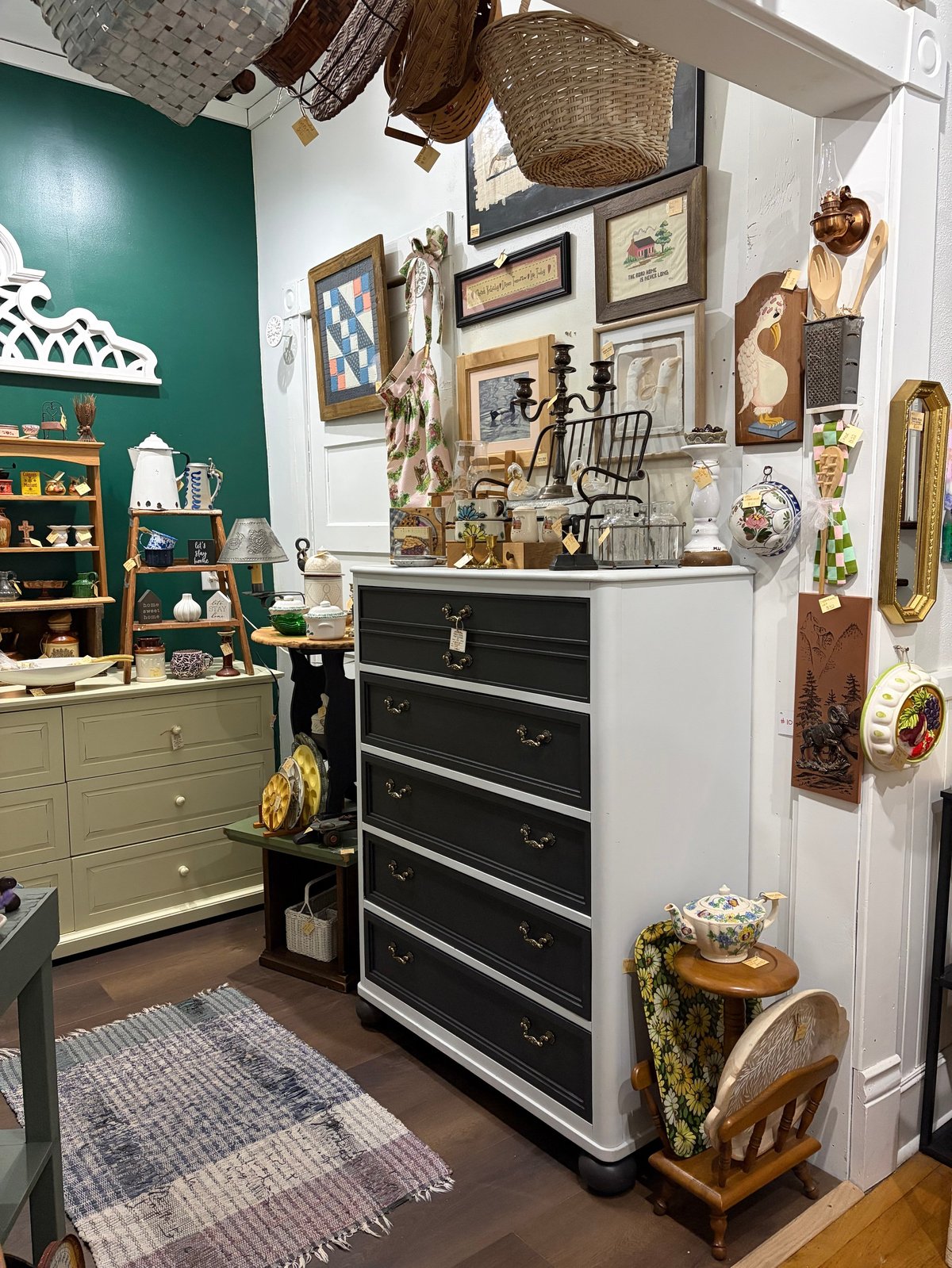 Eclectic vintage-inspired room with green accent wall, gallery wall art, black and white dresser, woven baskets, decorative mirrors, and various collectibles arranged throughout