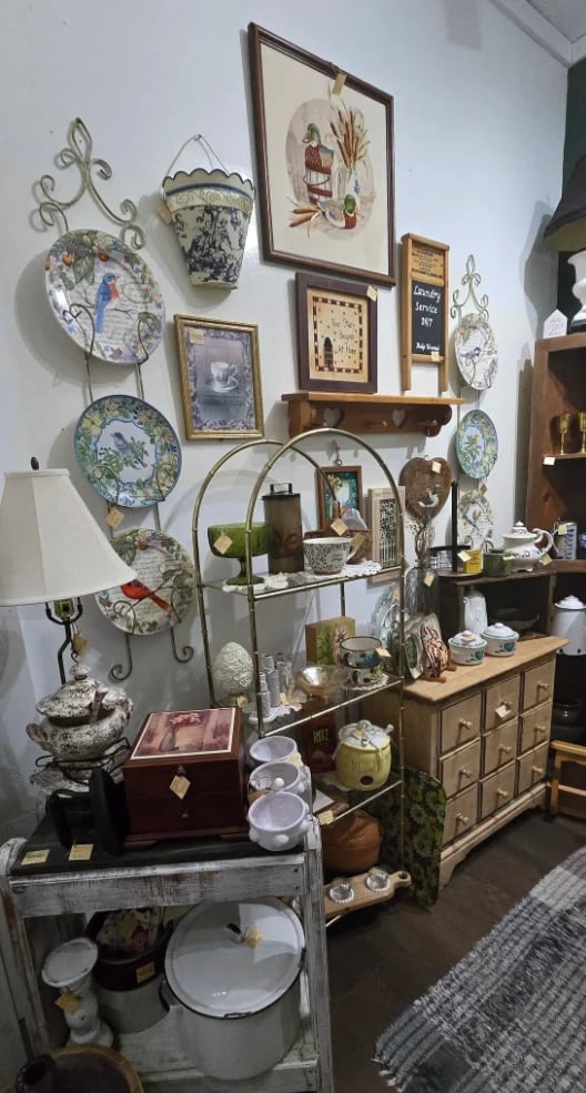Eclectic vintage decor display with decorative plates, shelves, lamp, framed prints, and antique furniture arrangements on white wall