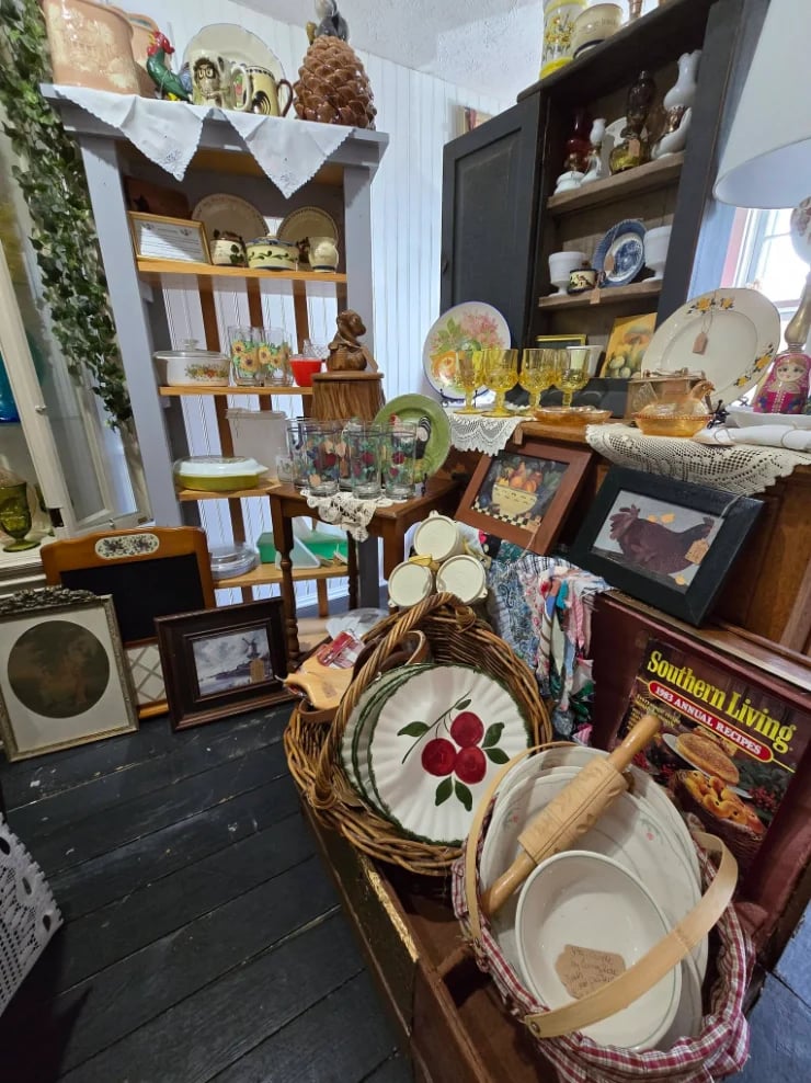 Antique shop interior with wooden shelves displaying vintage dinnerware, baskets of kitchen items, decorative plates, framed artwork, and country home decor arranged throughout the cluttered space
