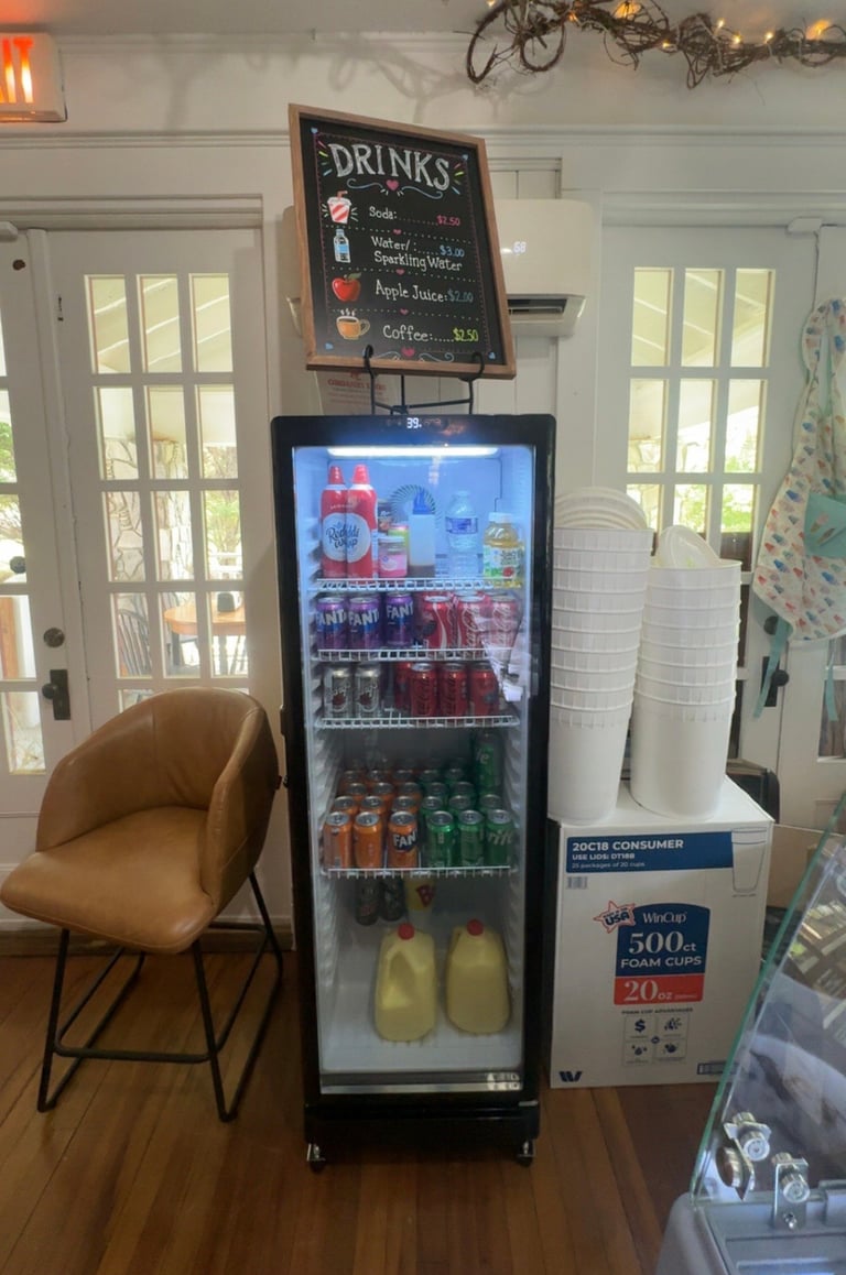 A refrigerated beverage cooler stocked with drinks and juices, with a chalkboard menu above it and a tan chair beside it in a bright café interior