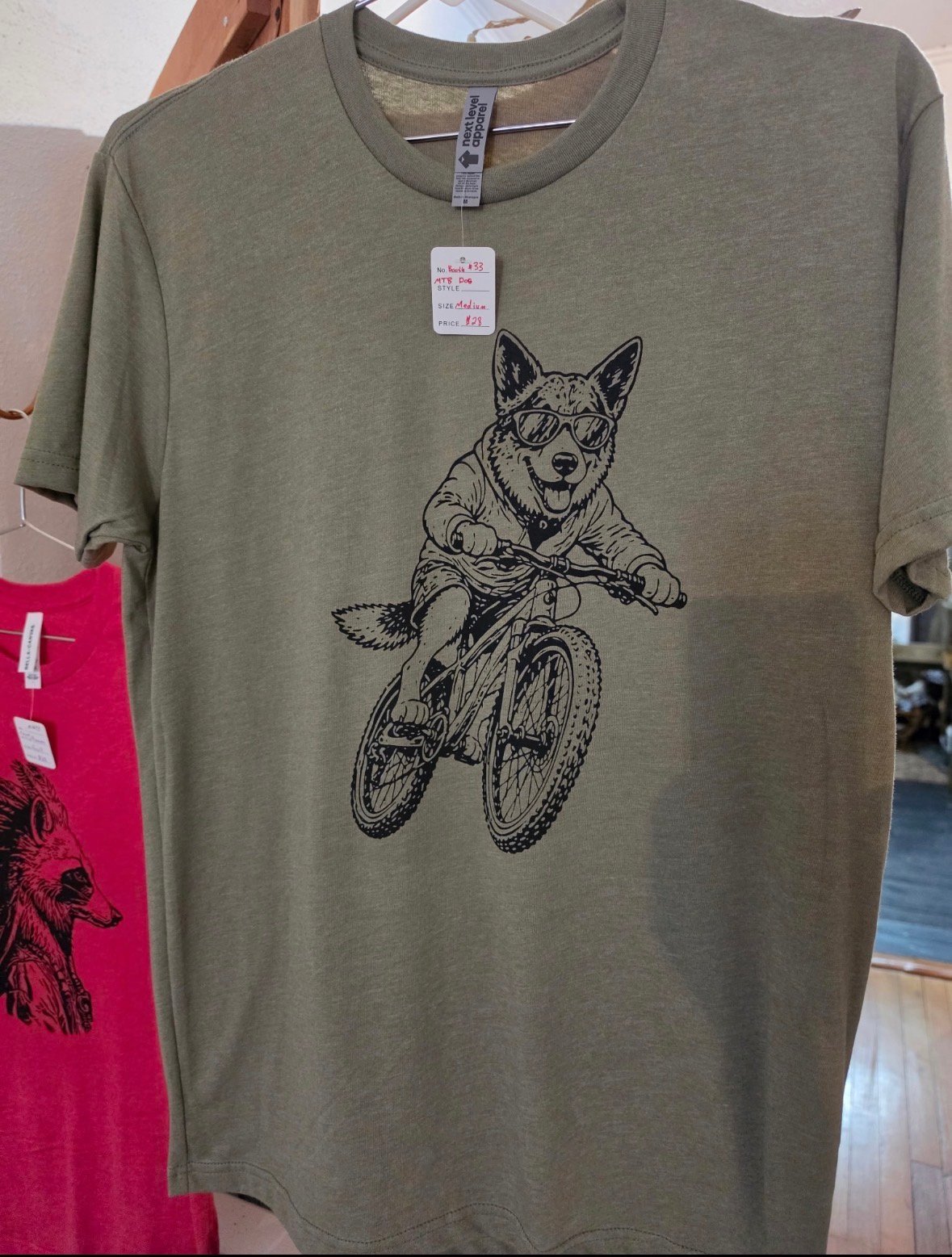 Sage green t-shirt with black illustration of a dog riding a bicycle