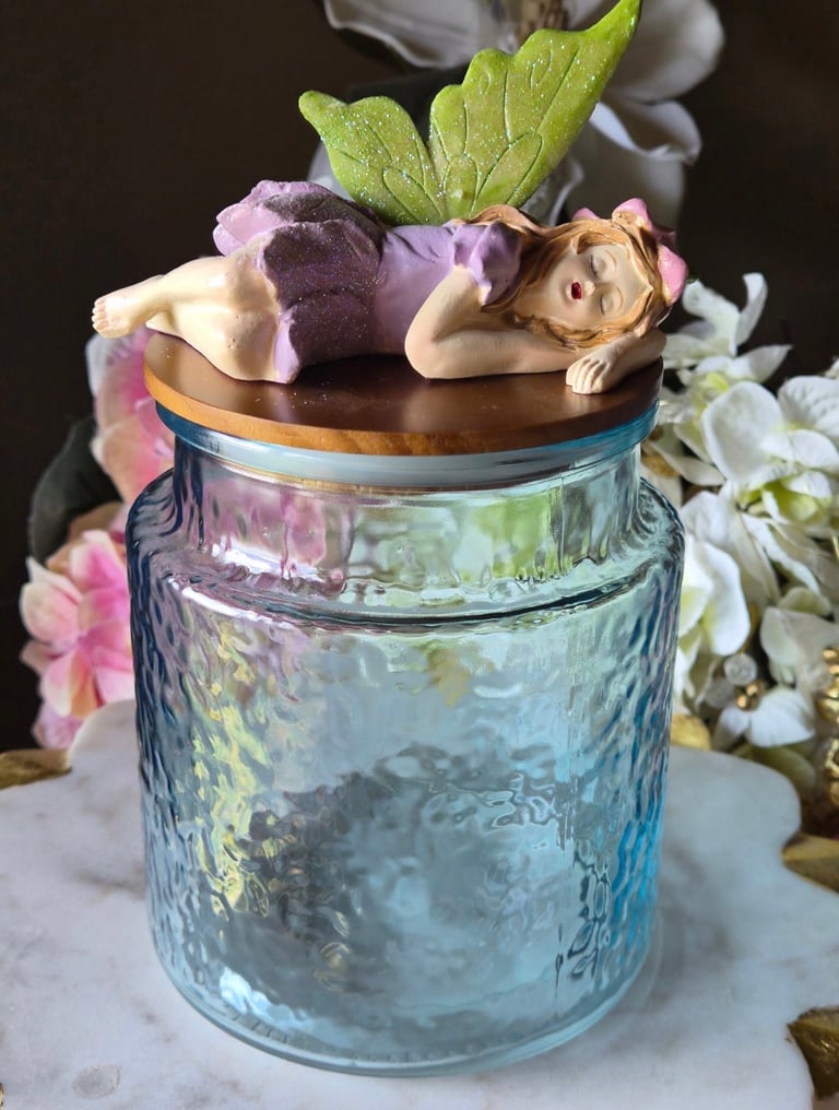 A decorative glass jar with a wooden lid topped with a cherub figurine, surrounded by pink and white flowers