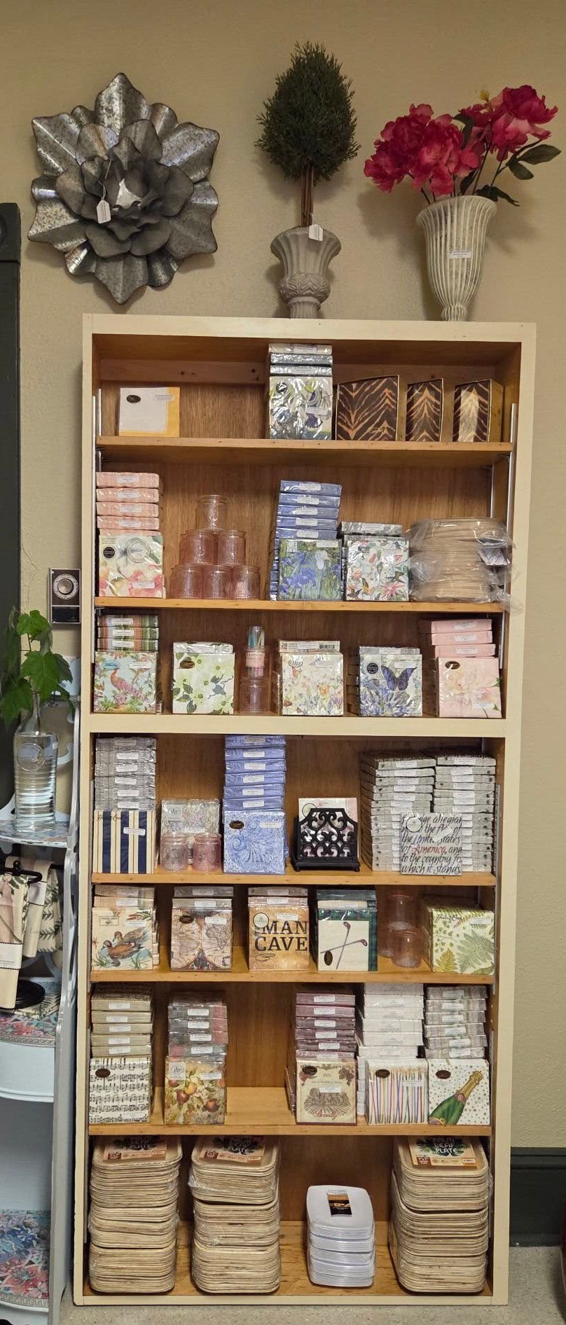 Organized wooden shelf unit displaying stacked decorative boxes and baskets, with silver bow, dried flowers, and plant decor on top against beige wall.