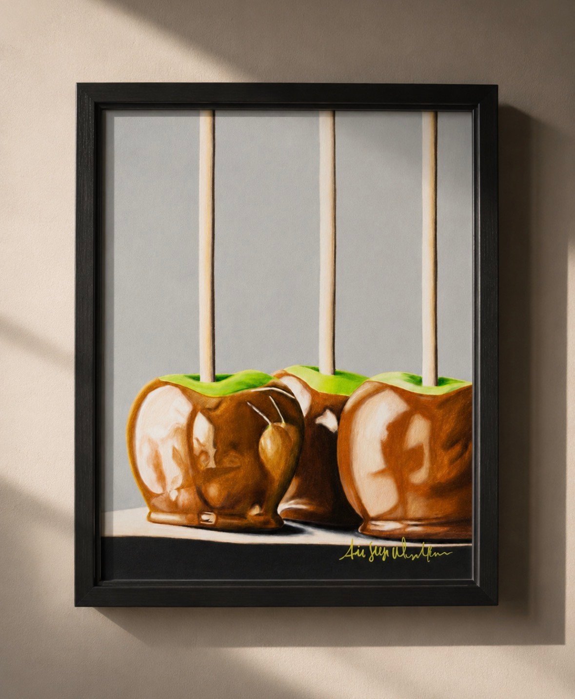 Framed artwork of three caramel apples with green leaf stems displayed in a black frame on a beige wall