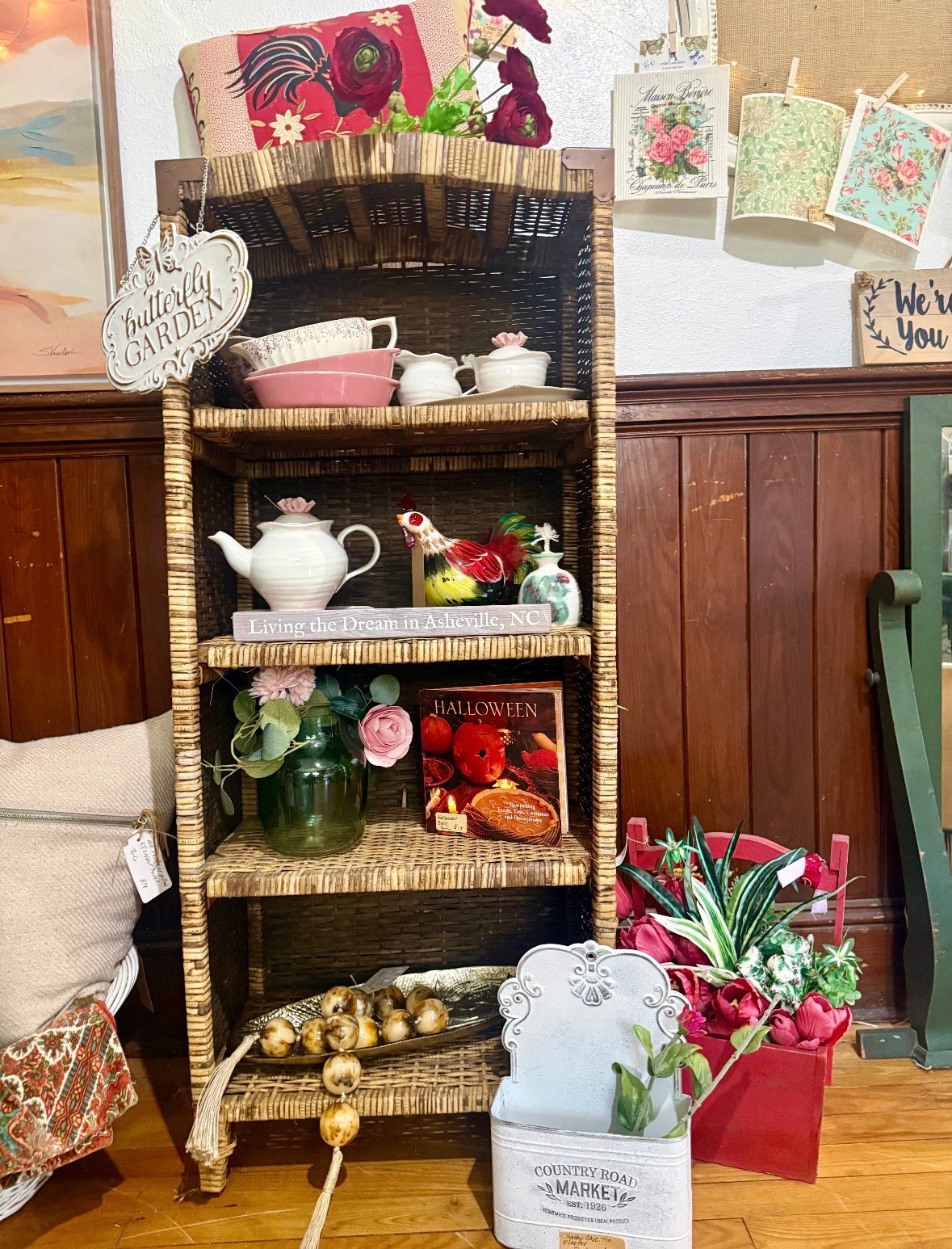 Woven wicker shelf unit displaying decorative items including teapot, flowers, floral artwork, and seasonal home décor
