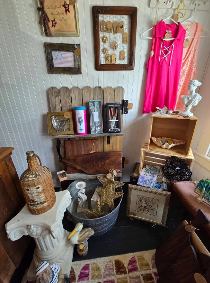Eclectic boutique display with rustic wooden shelving, bright pink dresses on hanger, woven baskets, decorative items, and various merchandise arranged on shelves and table