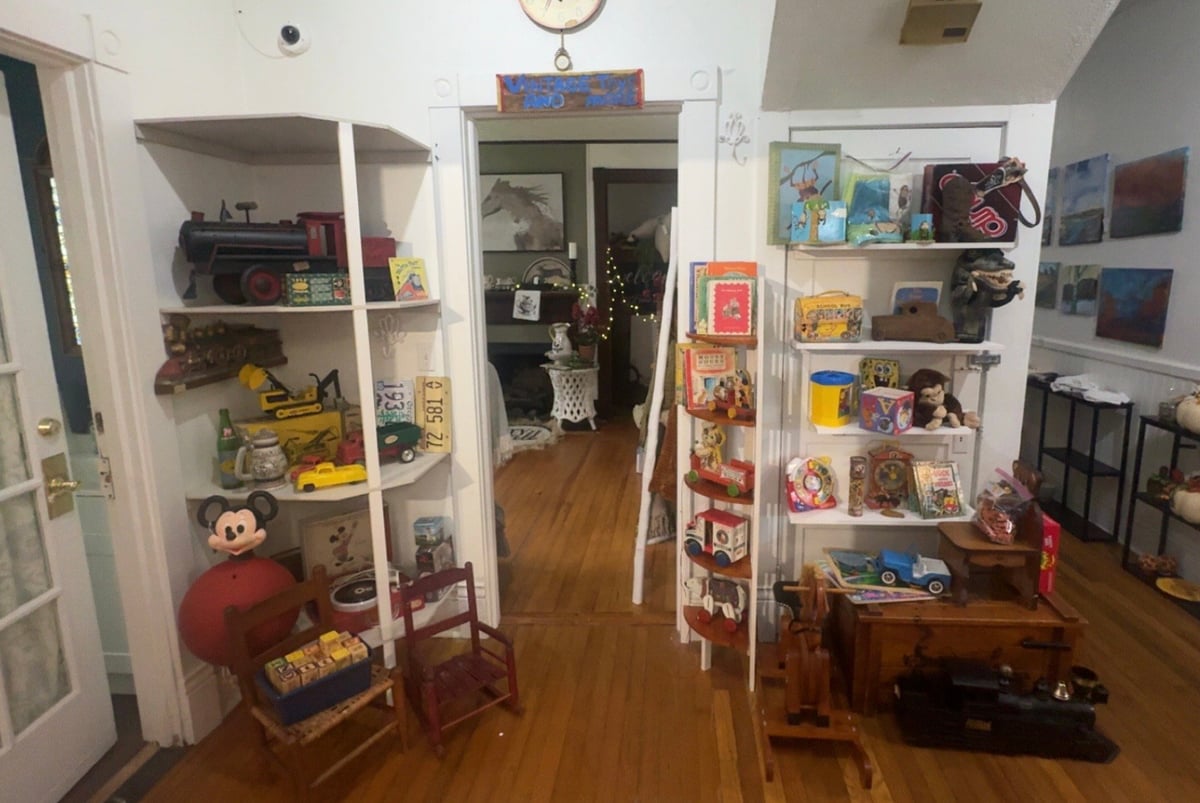 A museum or store display room with glass display cases containing collectibles, toys, and memorabilia on wooden shelves, with an open doorway visible in the background.