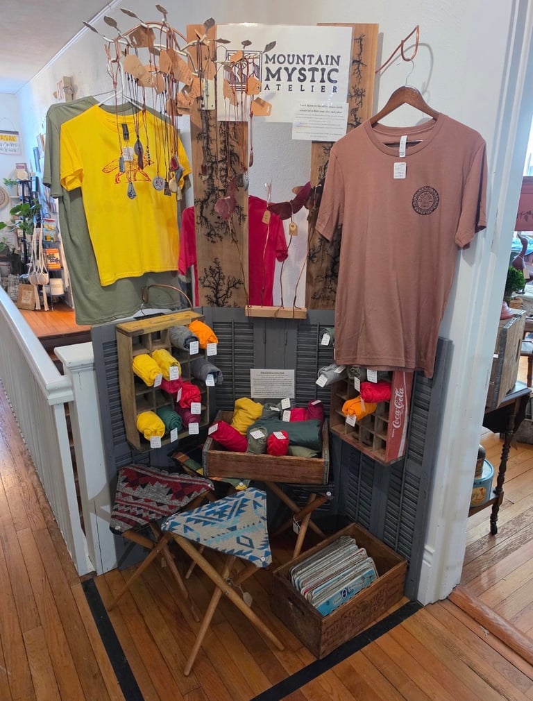 Retail display for Mountain Mystic Apparel featuring hanging clothing, folded items in bins, and textiles on wooden furniture