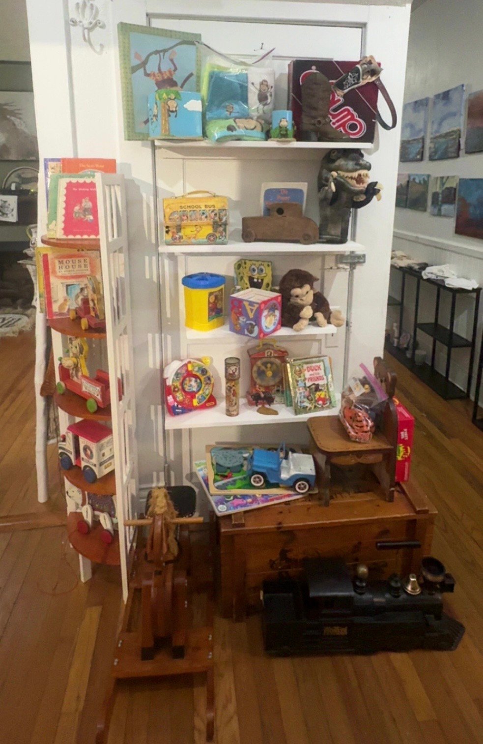 White shelving unit displaying a collection of vintage toys, lunch boxes, and collectibles on wooden floor