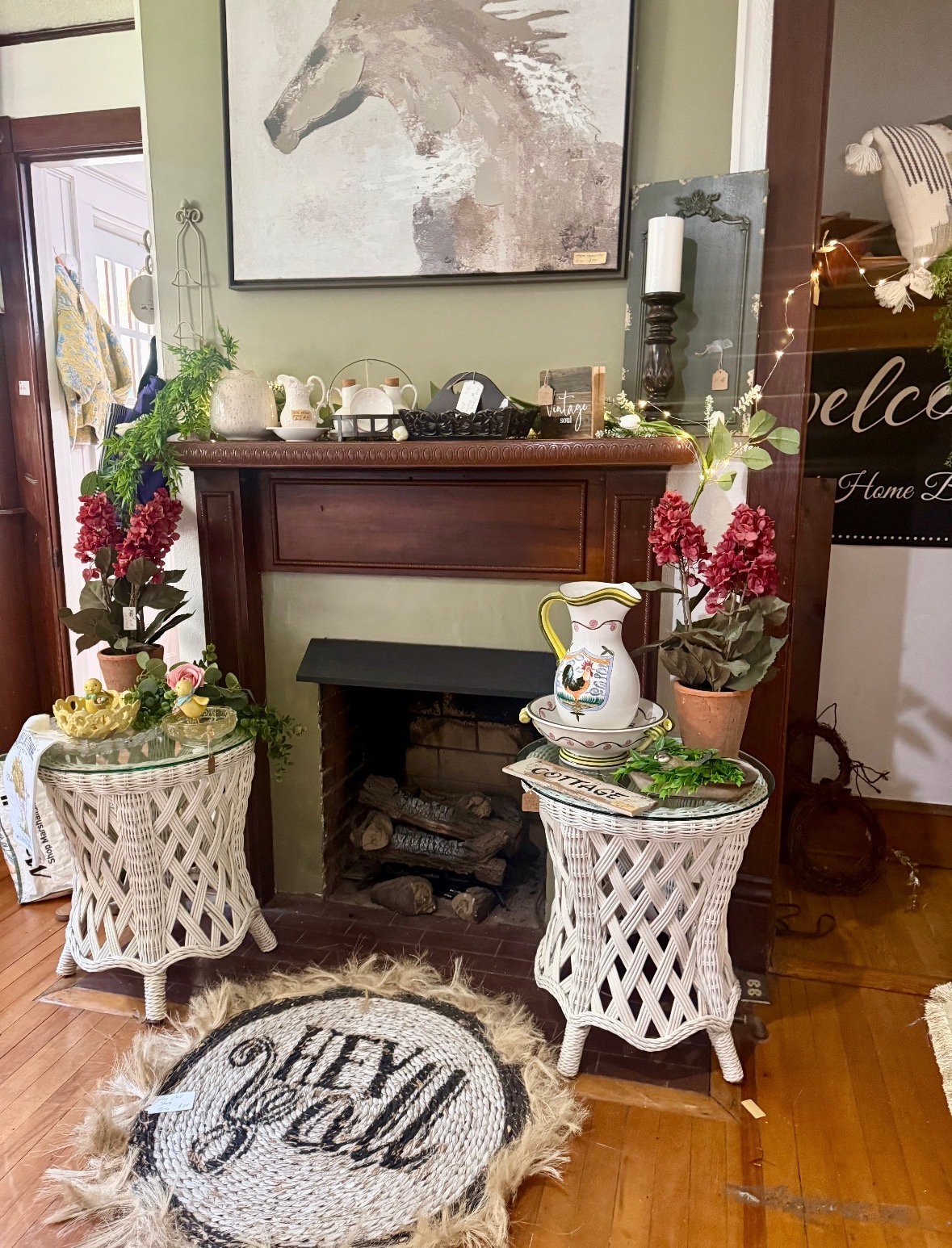 Decorated fireplace mantel with dark wood frame, flanked by white woven side tables with potted red flowers, horse portrait above, and welcome sign to the right