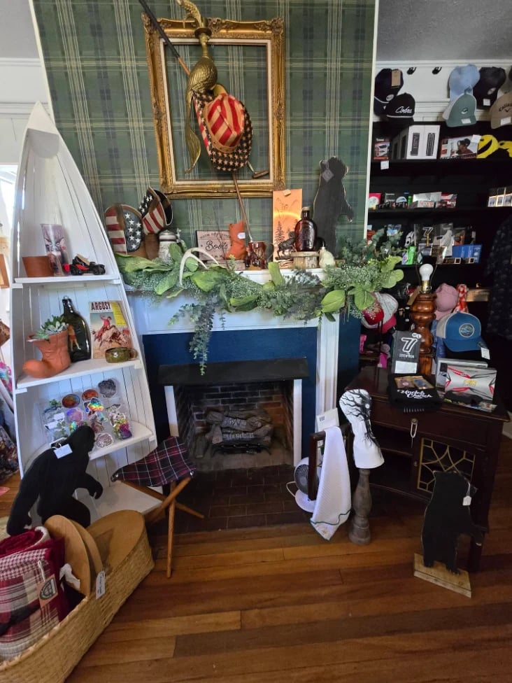 An eclectic living room display featuring a fireplace mantel decorated with plants, a gold-framed portrait above it, shelving units with collectibles, and various vintage decor items on wooden flooring