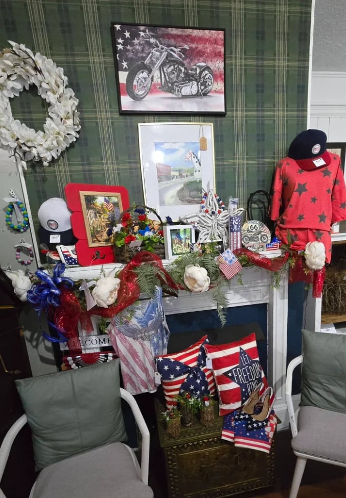 Patriotic memorial display with American flags, white flowers, framed photos, and red sweater on green striped wallpaper background