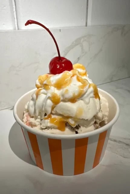 Vanilla ice cream sundae in striped cup topped with whipped cream, caramel drizzle, and a maraschino cherry on stem