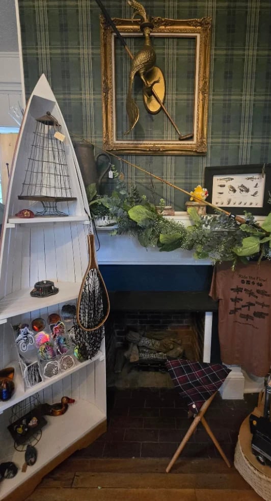 Fishing-themed home decor display with boat model, ornate gold frame with fish art, green plaid wallpaper, and fishing equipment on white shelving