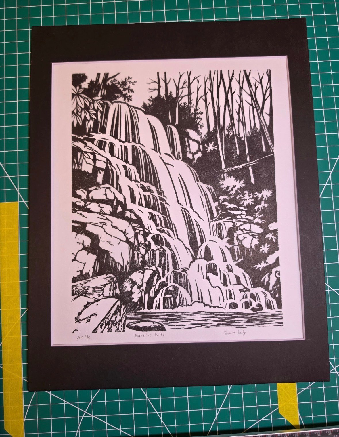 Black and white line drawing of a cascading waterfall in a forest with bare trees and rocks, mounted on a pink mat with dark border on a green cutting mat