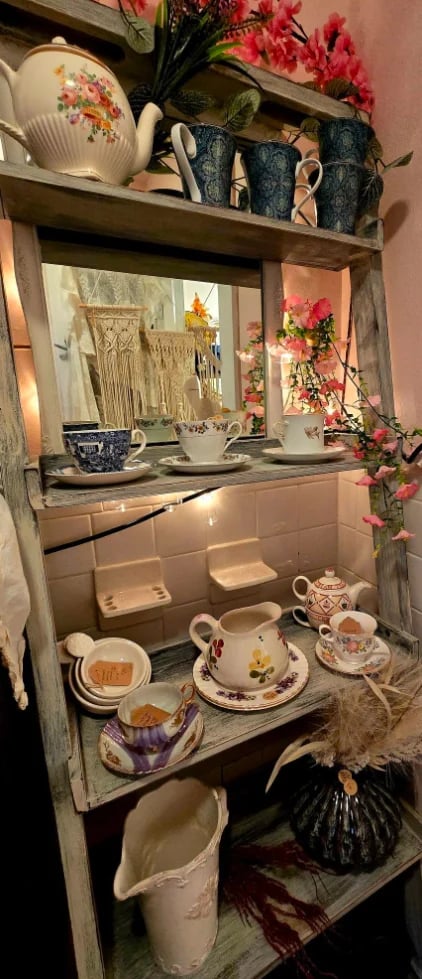 Wooden shelving unit displaying collection of decorative ceramic dishes, teacups, and floral arrangements against pink wall
