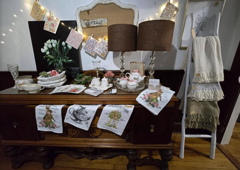 Vintage cottage-style display with wooden dresser, brown lampshades, decorative plates, potted plants, white linens, and hanging pendant lights