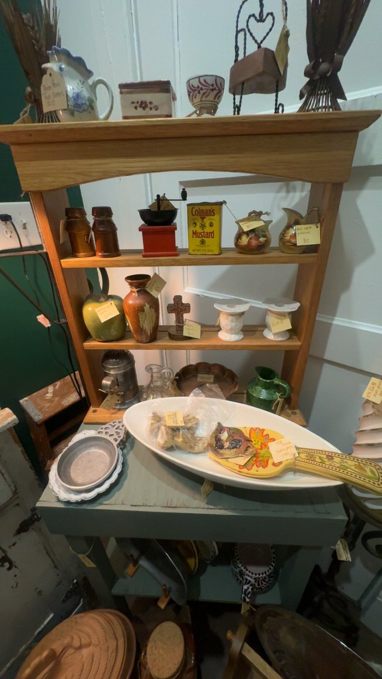 Wooden shelving unit displaying decorative items, vintage bottles, pottery, and plants with a table below holding dishware and food items