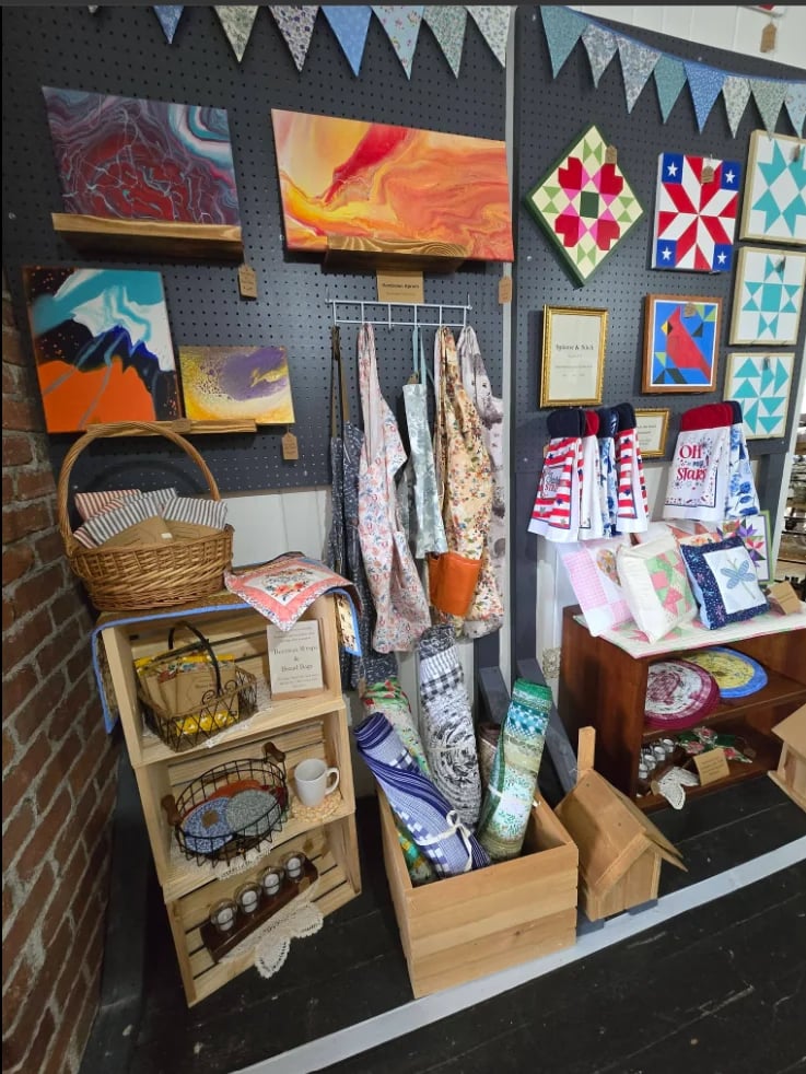 Retail display on pegboard wall featuring colorful artwork, bundles of fabric, woven baskets, wooden shelving with home goods, and decorative banners