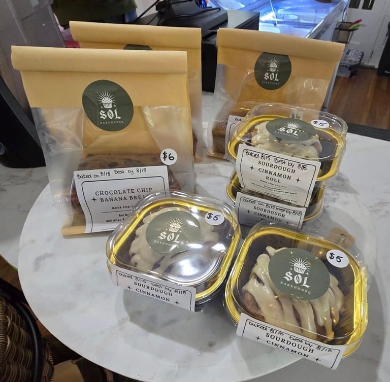 Assorted baked goods from SOL Bakehouse displayed on white marble surface, including kraft paper bags and clear containers with labeled items