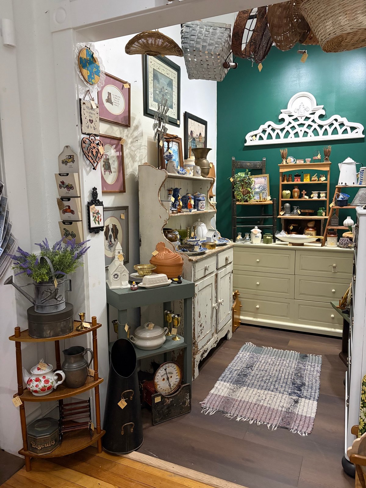 Eclectic vintage-style room with teal walls, decorative baskets, shelving units filled with collectibles, and a mix of farmhouse and antique decor.