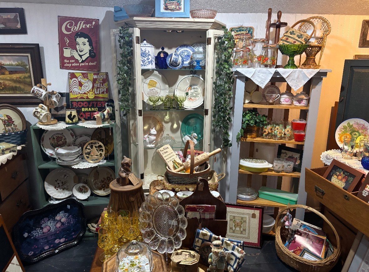 Antique shop display with vintage collectibles including decorative plates, glass items, figurines, and shelving units arranged throughout cluttered store.