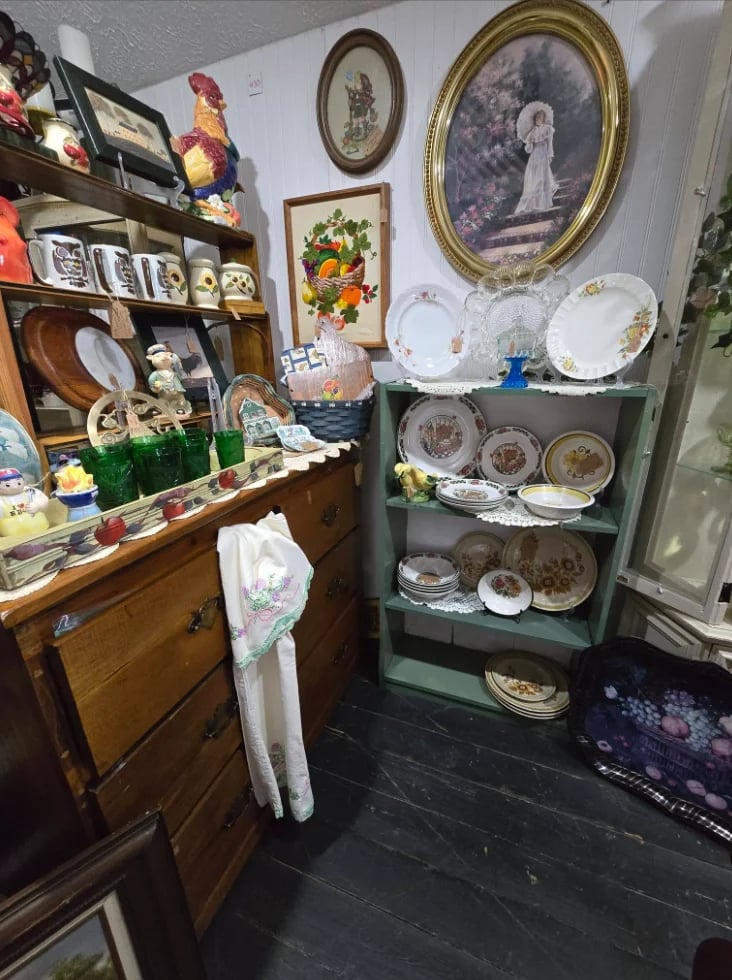 Antique display room with wooden furniture, framed portraits, decorative plates on green shelving unit, and various collectible items arranged throughout